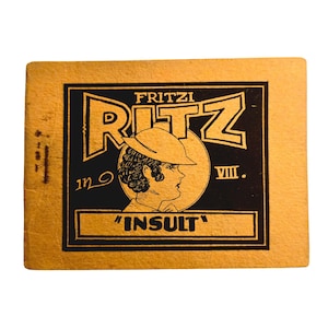 May include: A vintage Fritzi Ritz card with a black and gold design. The card features the name "Fritzi Ritz", a profile illustration of a woman wearing a hat, and the word "INSULT".