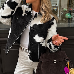 May include: A cow-print jacket with black and white patches, a white shirt, white trousers, and a dark brown suede handbag featuring a buckle strap and decorative charms. The jacket has a collar and black buttons down the front centre.