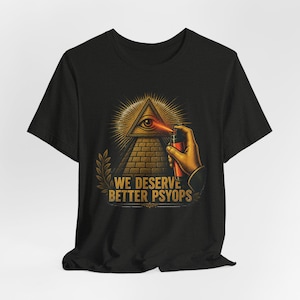 May include: Black t-shirt featuring a gold pyramid with an eye, a hand holding a red spray can, and the text "WE DESERVE BETTER PSYOPS" in gold. The design is a vintage-style illustration.