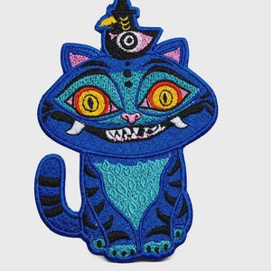 May include: An embroidered patch featuring a blue cat with large yellow eyes, a wide grin, and black stripes. A small black bird with a single eye sits on the cat's head. The design uses vibrant shades of blue, turquoise, yellow, and pink thread.