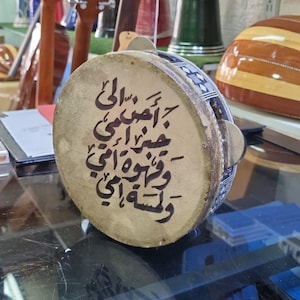 May include: A round, tan drum with Arabic calligraphy in black ink. The drum has a wooden frame and a patterned border. Guitars and other musical instruments are visible in the background.
