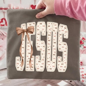 May include: A grey t-shirt with the word "JESUS" in a decorative font. The letters are filled with a pattern of small bows and accented with a large brown bow. The shirt is held by a person.