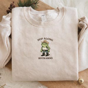 May include: A cream-colored sweatshirt with the text "Hop Along Buckaroo" and a cartoon frog wearing a cowboy hat, vest, and boots. The frog is green with a white vest and brown boots. The sweatshirt is folded on a wooden surface.