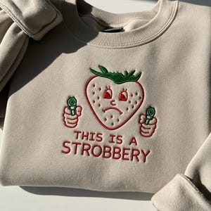 May include: Beige sweatshirt with an embroidered strawberry design. The strawberry has a sad face and is holding two green guns. The text "THIS IS A STROBBERY" is embroidered in red below the strawberry.