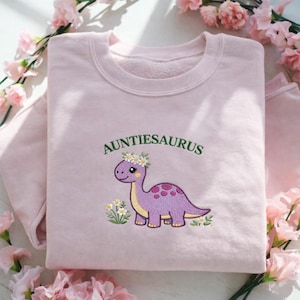 May include: A pale pink sweatshirt featuring the embroidered word "AUNTIESAURUS" above a purple dinosaur wearing a floral crown. The dinosaur has yellow details and is surrounded by embroidered flowers and foliage.