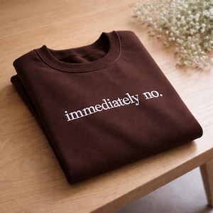 May include: A folded brown sweatshirt with the embroidered text "immediately no." The crew neck sweatshirt is made of a soft material and is folded neatly on a wooden surface. White flowers are in the background.