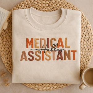 May include: A cream-coloured sweatshirt featuring the text "MEDICAL ASSISTANT" in a colourful, block letter design. The name "Halley" is written in a cursive font between the words. The sweatshirt is folded on a woven mat.