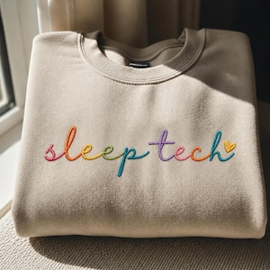 Peut inclure: Un sweat-shirt beige avec le texte brodé "sleep tech" en couleurs arc-en-ciel. Le sweat-shirt a un col rond et des manches longues. Le texte est en écriture cursive avec un petit cœur jaune à la fin.
