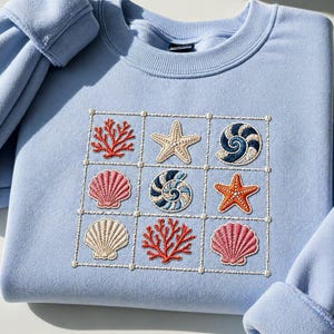 May include: A light blue sweatshirt featuring an embroidered square design with various sea creatures. The design includes a starfish, seashells, and coral in shades of orange, pink, and blue. The sweatshirt has long sleeves and a crew neck.