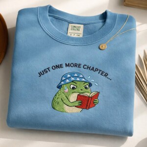 May include: Pale blue sweatshirt featuring an embroidered frog reading a book and wearing a bucket hat. The text "JUST ONE MORE CHAPTER..." is above the frog. A gold necklace is draped on the sweatshirt.