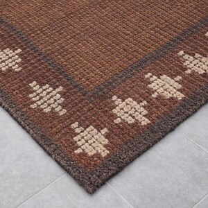 May include: A corner view of a brown rug with a geometric pattern. The rug has a dark brown border and a lighter brown center. The geometric pattern is a repeating design in a beige color.
