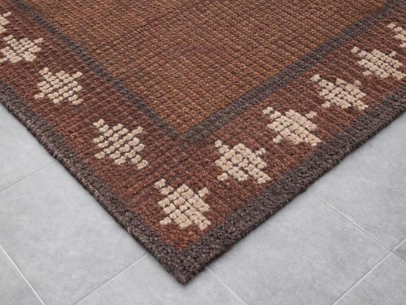 May include: A corner view of a brown rug with a geometric pattern. The rug has a dark brown border and a lighter brown center. The geometric pattern is a repeating design in a beige color.