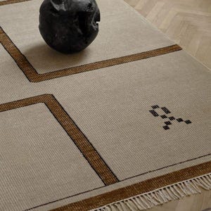 May include: Beige rectangular rug with brown geometric border and fringe. A dark, textured sphere sits on the rug. The rug has a simple, modern design, suitable for a living room or bedroom.