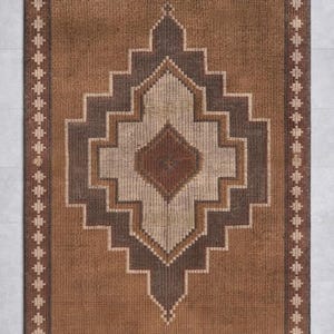 May include: A rectangular brown rug with a geometric diamond pattern in shades of brown and beige. The rug has a border with a repeating pattern of small, stylized shapes.