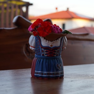 Op de afbeelding: Een keramische plantenbak in de vorm van een torso met een blauw-witte dirndl met rode veterdetails, een wit schort en pofmouwen. De opening aan de bovenzijde is gevuld met felrode geraniumbloemen voor een decoratieve uitstraling.