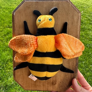 May include: A plush bee toy with yellow and black stripes, orange wings, and blue eyes is mounted on a wooden plaque. The bee has black antennae and legs. The wooden plaque is rectangular with rounded corners.