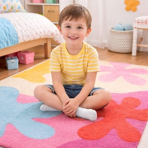 May include: A colorful, floral-patterned rug in a child's bedroom. The rug features large, abstract flower shapes in pink, blue, orange, and yellow. A child wearing a yellow striped shirt and denim shorts sits on the rug.