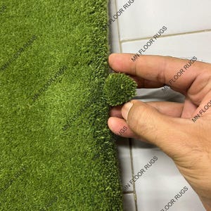 May include: Close-up of a vivid green rug with a dense, textured pile. A small, detached sample of the rug's material is held up, demonstrating the colour and texture. The rug appears to be a shag style, with a soft, inviting appearance. The text "MN FLOOR RUGS" is visible.