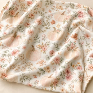May include: A white fabric featuring a repeating pattern of soft peach-coloured pumpkins, delicate pink flowers, and muted green foliage. The design includes scattered autumn leaves and small floral sprigs, creating a soft, light-toned textile print.