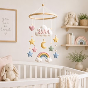 Pode incluir: Um móbile suspenso com uma estrutura circular de madeira e fios brancos que sustentam elementos de crochê, incluindo uma nuvem branca central bordada com o nome LUNA, uma lua crescente amarela, um arco-íris e estrelas em rosa, azul, amarelo e verde.