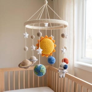 May include: A handmade crochet baby mobile featuring a solar system theme. The mobile includes a yellow sun, planets, stars, and an astronaut, all suspended from a cream-colored ring. The planets include Earth, Saturn, and other celestial bodies.