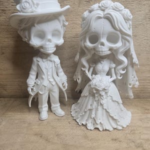 May include: A pair of white matte figurines depicting a skeletal couple. The figure on the left wears a top hat and suit with a rose boutonniere. The figure on the right wears a veil with roses and a gown, holding a bouquet of roses.