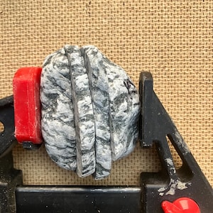 以下が含まれることがあります： 黒と赤のプラスチック製クランプでしっかりと固定された、3つの垂直なスライスにカットされたグレーと白のマーブル模様の石。石には自然な暗い筋模様があり、層状の構造全体に粗い質感の表面が見られます。