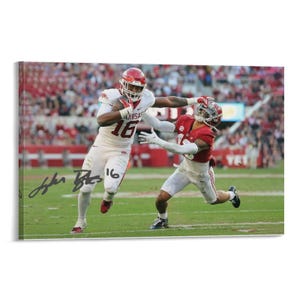 Könnte beinhalten: Gerahmtes Action-Foto eines Footballspiels. Ein Spieler in rotem Helm und Trikot, Nummer 16, läuft mit dem Ball, verfolgt von einem Spieler in rotem Trikot. Der Hintergrund zeigt ein Stadion voller Zuschauer.