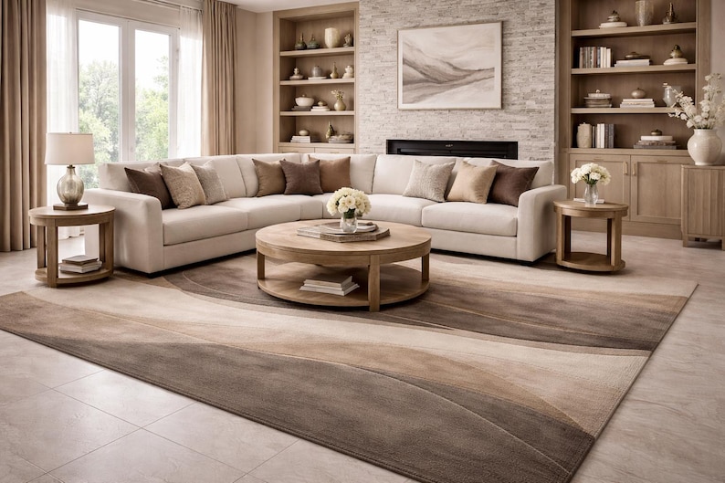 May include: A modern living room featuring a large, off-white sectional sofa with several throw pillows in shades of brown. A round wooden coffee table sits in front of the sofa, and a patterned rug in neutral tones covers the floor. The room has a fireplace and built-in shelves.