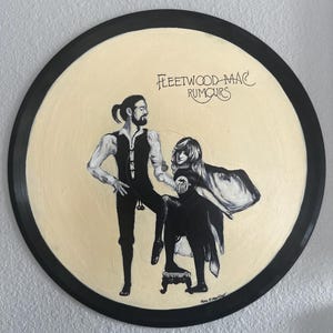 May include: A circular vinyl record painted with a cream-colored background featuring a black and white illustration of two figures. The text Fleetwood Mac Rumours is written in black script. One figure stands while the other sits on a stool.