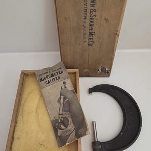 May include: A vintage Brown & Sharpe micrometer calliper with a dark metal frame and silver spindle, accompanied by its original wooden storage box and a printed instruction manual titled The Brown & Sharpe Micrometer Caliper.