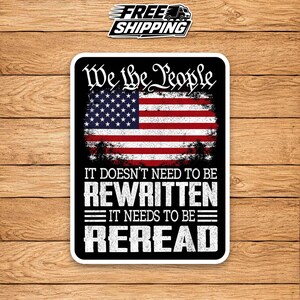 May include: A rectangular sticker with a black background featuring the American flag in the center. The text reads We the People at the top, and IT DOESN'T NEED TO BE REWRITTEN IT NEEDS TO BE REREAD in white, distressed, block lettering below the flag.