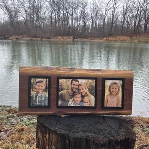 May include: A wooden picture frame with three black-framed photo openings. The frame is made from a natural wood slab and is displayed outdoors on a tree stump. The photos show a family and a child.
