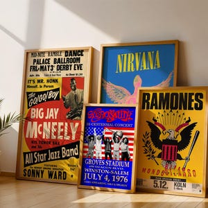 May include: A selection of framed concert posters with wooden frames. The posters display text and graphics for bands like Nirvana and Ramones. The posters are displayed against a neutral wall, with a potted plant in the corner.