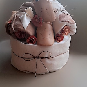 Könnte beinhalten: Ein Babygeschenkkorb mit hellbraunem Stoffboden, gebunden mit einem braunen Band. Der Korb enthält Babyschuhe, eine Decke und Kleidung in Braun- und Weißtönen, verziert mit roten Rosen.