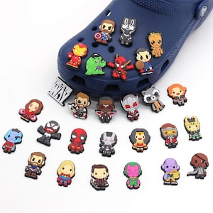 May include: A collection of colorful cartoon character shoe charms, including Iron Man, Hulk, and Deadpool, displayed on a blue shoe. The charms are various sizes and shapes, with detailed designs. The text "THIS IS THE WAY" is also visible.