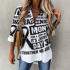 Op de afbeelding: Witte button-down shirt met zwarte tekst en een zwart lintontwerp. De tekst bevat de woorden "NATIONAL AWARENESS MONTH" en "FIND A CURE BUT IT'S EVERY DAY 3". De shirt heeft een V-hals en driekwart mouwen.