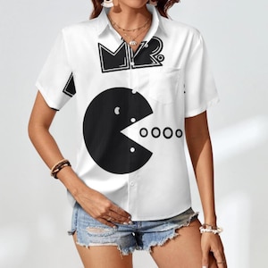 Op de afbeelding: Wit overhemd met een grote zwarte Pac-Man-afbeelding. Het shirt heeft korte mouwen, een kraag en een borstzak. De bovenkant van het shirt heeft de letters "MR" in zwart.