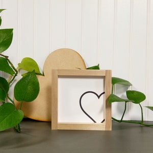 May include: A square wooden framed sign with a black heart outline on a white background. A large wooden heart shape is behind the sign. Green leafy plants are in a metal container and trailing across the surface.