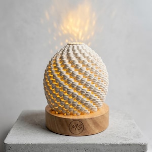 May include: A cream-coloured, spherical essential oil diffuser with a textured, woven design. The diffuser sits on a wooden base with the initials "MS" and emits a warm, glowing light. The product is placed on a grey surface.
