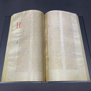 May include: Open book with aged, yellowed pages. Text is in columns with red and black lettering. The letter "H" is in red at the beginning of a paragraph. The book is open on a dark surface.