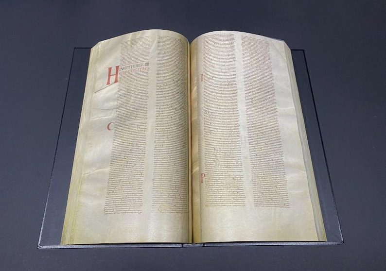 May include: Open book with aged, yellowed pages. Text is in columns with red and black lettering. The letter "H" is in red at the beginning of a paragraph. The book is open on a dark surface.