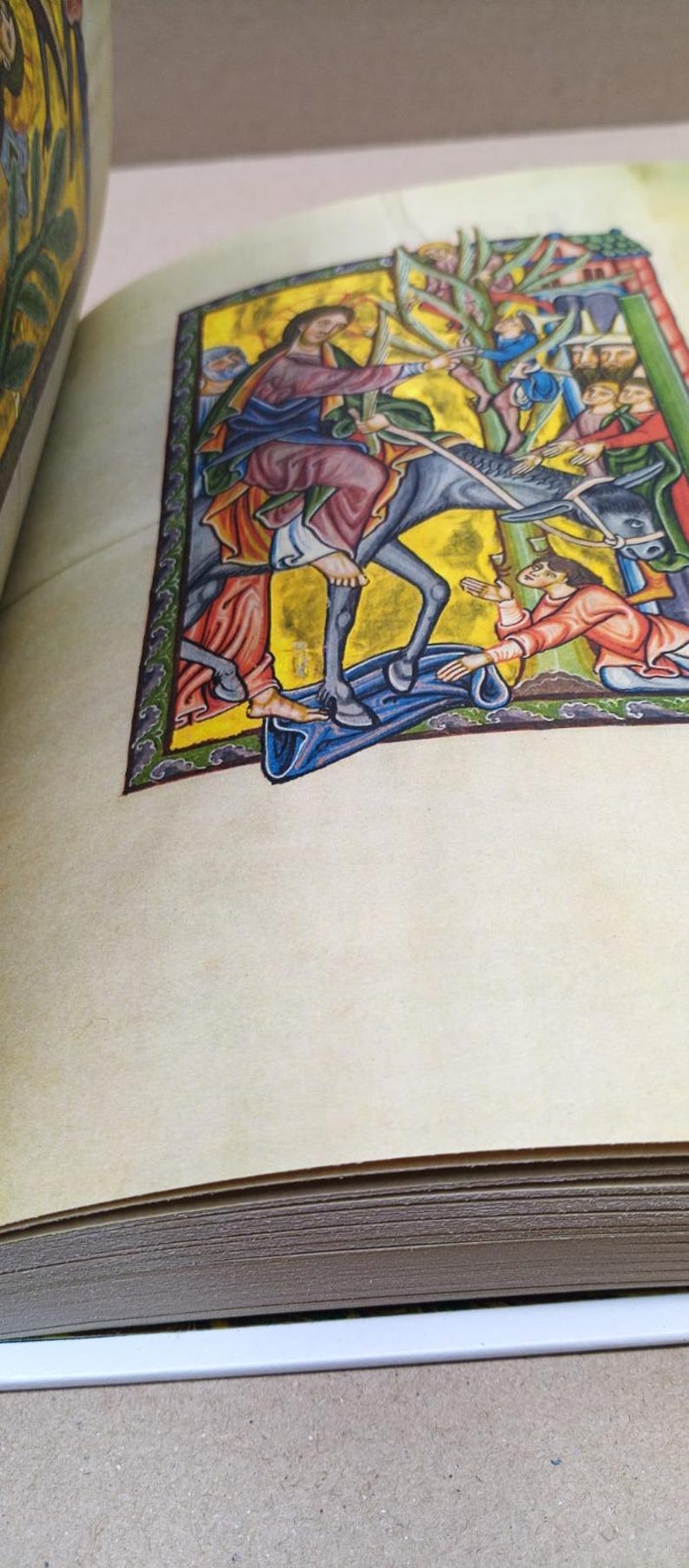 Może przedstawiać: Otwarta książka prezentująca żywą średniowieczną ilustrację. Grafika przedstawia postać jadącą na osiołku, otoczoną przez ludzi w kolorowych szatach. Scena rozgrywa się na złotym tle ze szczeg&oacute;łowymi obramowaniami.