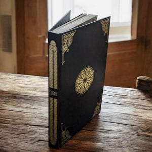 May include: A black book with gold decorative accents and the text "Codex Gigas" on the spine. The book has a gold circular design on the cover and is open to reveal the pages. The book is on a wooden surface.