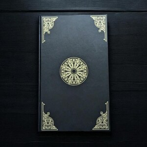 May include: A black journal with gold decorative accents. The cover features a central circular design and ornate corner details. The journal is rectangular and appears to be closed, resting on a dark surface.