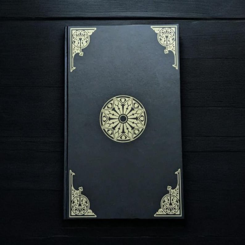 May include: A black journal with gold decorative accents. The cover features a central circular design and ornate corner details. The journal is rectangular and appears to be closed, resting on a dark surface.
