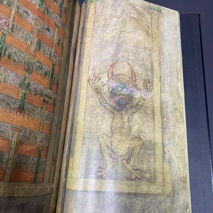 May include: Open book with a detailed illustration of a horned, green-faced creature with outstretched arms. The creature is depicted in a seated position, with a yellow border around the image. The opposite page shows a detailed architectural drawing.