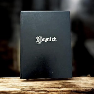 May include: A black book with the word "Voynich" in white gothic lettering. The book is standing upright on a piece of wood. The background is dark and out of focus.