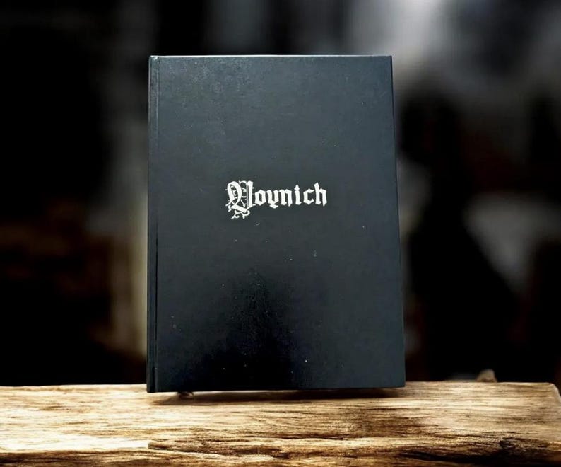 May include: A black book with the word "Voynich" in white gothic lettering. The book is standing upright on a piece of wood. The background is dark and out of focus.