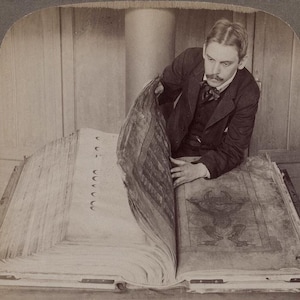May include: A sepia-toned photograph of a man examining a large, open book. The book's pages are thick and aged, with illustrations and text visible. The man is wearing a dark suit and bow tie, engrossed in the book's contents.